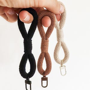 Puede incluir: Tres llaveros tejidos en negro, marrón y beige. Cada llavero presenta un diseño de bucle con un cierre metálico. Los llaveros se sostienen sobre un fondo blanco, mostrando sus diferentes colores y texturas.