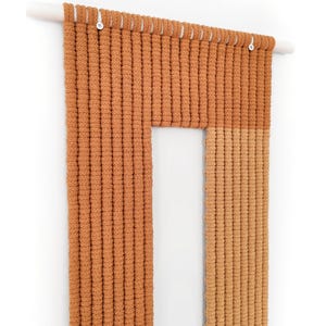 May include: A woven wall hanging with a rectangular shape and a smaller rectangular opening in the centre. The hanging is made of thick, brown yarn and is attached to a white wooden dowel rod.