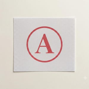 Puede incluir: Un lienzo de punto de aguja blanco que presenta un borde circular rojo con una letra A mayúscula roja grande centrada en el interior. El lienzo tiene una cuadrícula uniforme de pequeños agujeros en toda la superficie para bordar.