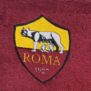 Può includere: Asciugamano bordeaux con lo stemma della squadra AS Roma ricamato. Lo stemma è a forma di scudo con sfondo giallo, una lupa bianca e grigia che allatta due bambini e la scritta "ROMA 1927" in oro.