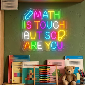 Może przedstawiać: Wielokolorowy neon z napisem MATH IS TOUGH BUT SO ARE YOU, zawierający ikonę ołówka i żarówki. Neon jest zamontowany na zielonej ścianie nad drewnianym biurkiem, na którym znajdują się książki, liczydło, pluszowy miś oraz kolorowe kredki.