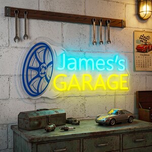 Könnte beinhalten: Ein Neonschild mit der Aufschrift "James's GARAGE" in Blau und Gelb, mit einem blauen Autorad-Design. Das Schild befindet sich an einer Backsteinmauer über einer Metallwerkzeugkiste, einem Modellauto und Werkzeugen.