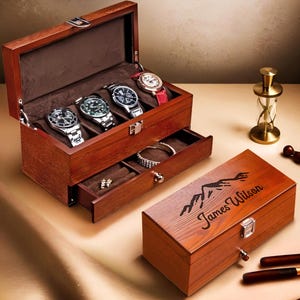 May include: A wooden watch box with a hinged lid, containing four watches with silver and black faces and a red leather strap. A drawer holds jewellery. The box is engraved with "James Wilson" and a mountain design.