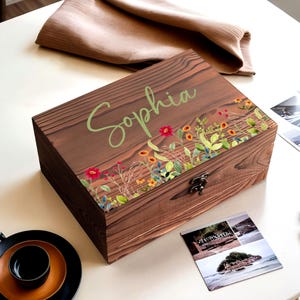 Puede incluir: Una caja de madera marrón oscuro con el nombre "Sophia" en verde. La caja está decorada con un borde floral de flores rojas, amarillas y azules. Un cierre de metal asegura la caja. Ideal para guardar cosas.