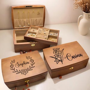 May include: Wooden jewelry boxes in various sizes and designs. Some boxes are open, revealing compartments for rings, necklaces, and bracelets. Two boxes have floral and name engravings. The boxes are made of light brown wood.