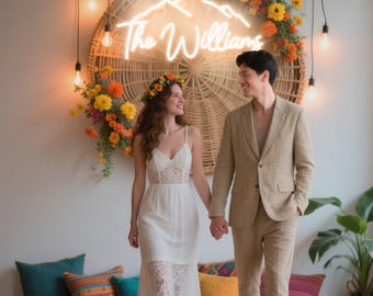 May include: A couple holding hands, smiling, in front of a wall with a neon sign that says "The Williams". The sign is above a woven circular decoration with flowers. The woman wears a white dress and flower crown. The man wears a tan suit.