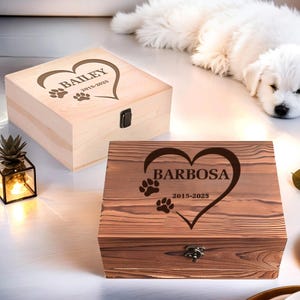 Op de afbeelding: Twee houten gedenkdozen voor huisdieren. Eén is lichtgekleurd met "Bailey" en pootafdrukken in een hart. De andere is donker gebeitst met "Barbosa" en pootafdrukken in een hart. Beide dozen hebben data en metalen sluitingen.