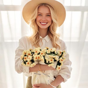 May include: A woman wearing a straw hat and white shirt holds a bouquet of crocheted flowers. The flowers are yellow with orange centers and green leaves. The bouquet is wrapped in white fabric with a ribbon that says "Happy".