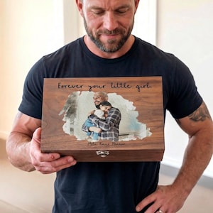 Puede incluir: Una caja de recuerdos de madera con una fotografía de un padre y un niño abrazándose. La caja está grabada con las palabras "Forever your little girl". La caja la sostiene una persona que lleva una camiseta negra.