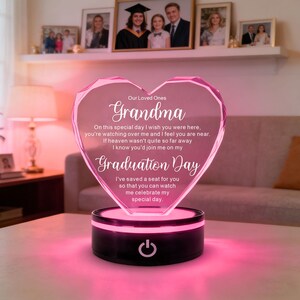 Puede incluir: Una luz acrílica en forma de corazón, teñida de rosa, con las palabras "Abuela" y "Día de Graduación" inscritas. La luz se encuentra sobre una base negra con un botón de encendido.