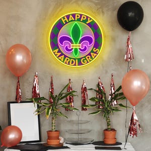 May include: A festive Mardi Gras scene with a neon sign, balloons, and plants. The sign reads "Happy Mardi Gras" with a fleur-de-lis symbol. Balloons are rose gold and black. Plants in terracotta pots and decorative tassels complete the party decor.