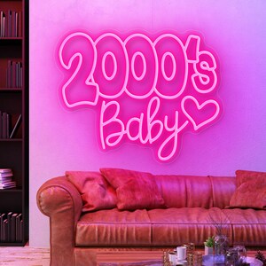 May include: A neon sign in the shape of the words "2000's Baby" in a pink hue, with a heart symbol. The sign is mounted on a pink wall above a brown leather sofa with red pillows. A bookshelf is visible on the left.
