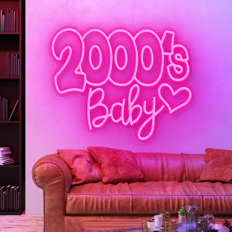 May include: A neon sign in the shape of the words "2000's Baby" in a pink hue, with a heart symbol. The sign is mounted on a pink wall above a brown leather sofa with red pillows. A bookshelf is visible on the left.