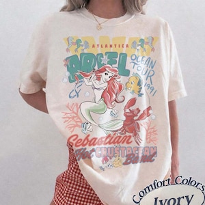 May include: Ivory-colored t-shirt featuring a graphic of Ariel from The Little Mermaid, with the text "Ocean Tour 1991." The design includes illustrations of Ariel, Flounder, and Sebastian. The shirt is labeled "Comfort Colors."