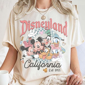 May include: Cream-colored t-shirt featuring a Disneyland California graphic with Mickey Mouse, Minnie Mouse, Donald Duck, Goofy, and Pluto. The graphic is in a circle with a pink and blue checkered background. The text "Est 1955" is below the word "California".