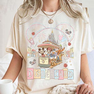 May include: Cream-colored t-shirt featuring a Disneyland design. The graphic shows Mickey Mouse, Minnie Mouse, Donald Duck, and Goofy riding a Main Street Trolley. The word "Disneyland" is written in pastel colors below the trolley.