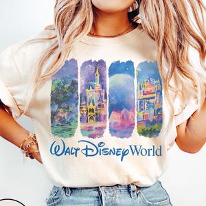 May include: Ivory-colored t-shirt featuring a colorful graphic of Walt Disney World landmarks. The design includes illustrations of a castle, fireworks, and other park features. The words "Walt Disney World" are printed in blue.