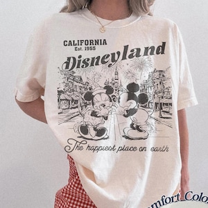 Puede incluir: Camiseta de color marfil con un gr&aacute;fico de Disneyland con Mickey y Minnie Mouse. El gr&aacute;fico incluye el texto "CALIFORNIA Est. 1955" y "The happiest place on earth". La camiseta se combina con pantalones cortos a cuadros rojos y blancos.