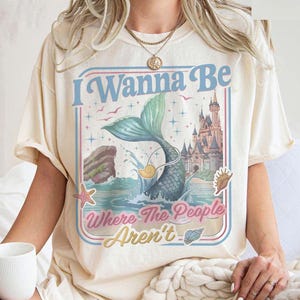 May include: Cream-colored t-shirt with a mermaid graphic and the text "I Wanna Be Where The People Aren't." The design features a mermaid tail, castle, and ocean elements in blue, pink, and yellow. The shirt has short sleeves.