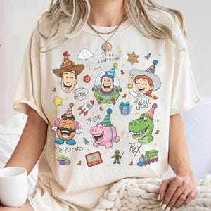 May include: Cream-colored t-shirt featuring colorful cartoon illustrations of characters from the Toy Story movie. The design includes Woody, Buzz Lightyear, Jessie, Mr. Potato Head, Rex, and Hamm, with the text "Buzz Lightyear".
