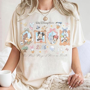 May include: Cream-colored t-shirt featuring a colorful graphic of Disney characters in framed portraits. The design includes Mickey Mouse, Minnie Mouse, Donald Duck, and Goofy, with the text "Walt Disney World" and "2026". The shirt also has the phrase "The Most Magical Place on Earth."