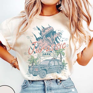 Può includere: T-shirt color avorio con grafica retrò "Jurassic Park". Il design presenta un dinosauro, un'auto e palme. Le parole "Jurassic Park" sono in un font stilizzato. La maglietta è realizzata in materiale morbido.