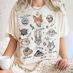 Może przedstawiać: Kremowa koszulka z portretami postaci z kresk&oacute;wki z filmu Zwierzogr&oacute;d. Postacie to m.in. Nick Wilde, Judy Hopps i Flash, z imionami takimi jak Gary De'Snake i Mr. Big. Koszulka ma swobodny, obszerny kr&oacute;j.