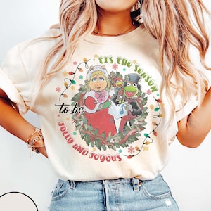 Puede incluir: Camiseta de color marfil con un diseño festivo con Miss Piggy, Kermit la Rana y Fozzie Bear cantando villancicos. El diseño incluye el texto "'Tis the season to be jolly and joyous" en una corona circular con luces navideñas.