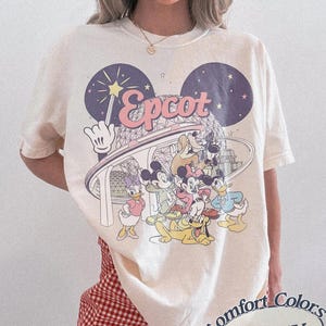 Peut inclure: T-shirt ivoire avec un motif de personnages Disney et le mot "Epcot" en rose. Le design comprend Mickey Mouse, Minnie Mouse, Donald Duck, Daisy Duck, Pluto et Tic et Tac. Le t-shirt est étiqueté "Comfort Colors Ivory".