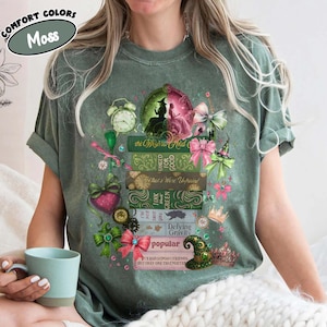 以下が含まれることがあります： モスグリーンのComfort Colors Tシャツ。デザインは、「The Wizard and」、「Defying Gravity」、「Popular」などのタイトルが書かれた本の山です。イラスト、リボン、魔女の帽子が含まれています。ゆったりとしたフィット感のTシャツです。