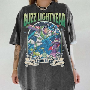 Puede incluir: Camiseta gris oscuro con un gráfico colorido de Buzz Lightyear de Toy Story. El diseño incluye los textos "Buzz Lightyear", "Disney", "Toy Story" y "Laser Blast". La camiseta tiene mangas cortas y un corte holgado.