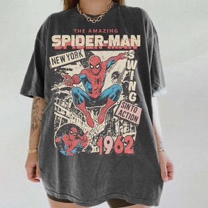 K&ouml;nnte beinhalten: Dunkelgraues T-Shirt mit einem Vintage-Comic-Design von Spider-Man. Die Grafik enth&auml;lt den Text "The Amazing Spider-Man", "New York" und das Jahr "1962". Der Superheld ist in seinem klassischen rot-blauen Anzug dargestellt.