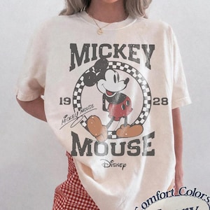 May include: Ivory-colored t-shirt featuring a distressed graphic of Mickey Mouse. The design includes the words "Mickey Mouse" and "Disney" with the years "1928" and "19". The shirt is a Comfort Colors brand.