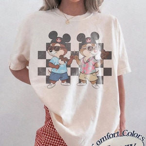 May include: Ivory-colored t-shirt featuring a graphic of two cartoon chipmunks wearing sunglasses and Mickey Mouse hats, holding ice cream cones. The chipmunks stand in front of a black and white checkered background. The shirt has the words "Comfort Colors Ivory" printed on it.