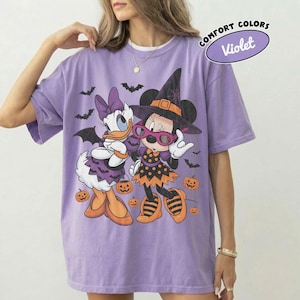 Może przedstawiać: Jasnofioletowy t-shirt z kr&oacute;tkim rękawem z grafiką przedstawiającą Daisy Duck i Minnie Mouse w kostiumach na Halloween. Wz&oacute;r zawiera nietoperze, pomarańczowe dynie oraz napis Comfort Colors Violet. Koszulka ma okrągły dekolt.