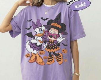 May include: A light purple short-sleeve t-shirt featuring a graphic of Daisy Duck and Minnie Mouse dressed in Halloween costumes. The design includes bats, orange pumpkins, and the text Comfort Colors Violet. The shirt has a crew neckline.