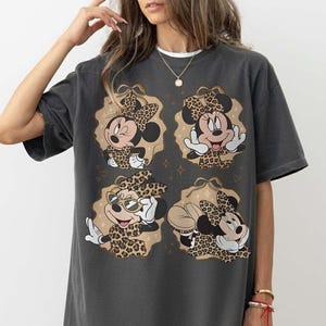 May include: Dark gray t-shirt featuring four images of Minnie Mouse in leopard print outfits. Each image is set within a tan oval with gold star accents. Minnie is wearing a bow and various poses. The shirt is a casual, oversized style.