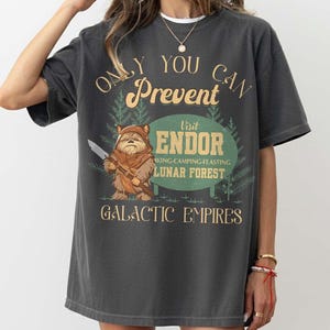 Puede incluir: Camiseta gris oscuro con un diseño gráfico de estilo vintage. El diseño presenta un personaje Ewok sosteniendo un cuchillo, con el texto "Only You Can Prevent Visit Endor Lunar Forest Galactic Empires."