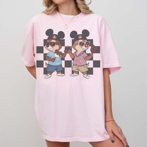 May include: A light pink t-shirt featuring a cartoon graphic of two chipmunks wearing sunglasses and Mickey Mouse ears. The chipmunks are holding ice cream cones and standing in front of a black and white checkered background.