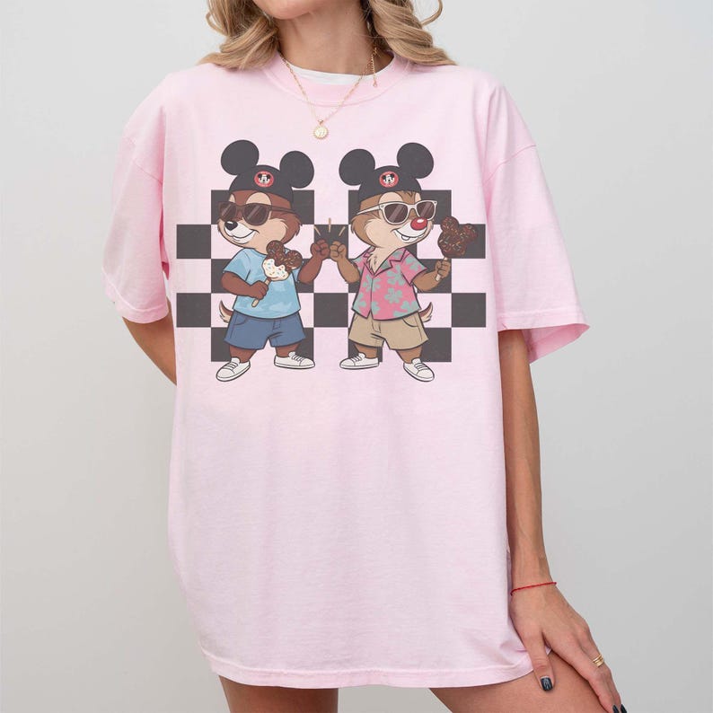 May include: A light pink t-shirt featuring a cartoon graphic of two chipmunks wearing sunglasses and Mickey Mouse ears. The chipmunks are holding ice cream cones and standing in front of a black and white checkered background.