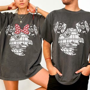 Puede incluir: Dos camisetas gris carbón con un diseño gráfico blanco. Una camiseta tiene un lazo de Minnie Mouse, la otra no. El diseño combina elementos de la Estrella de la Muerte y cazas TIE de Star Wars.