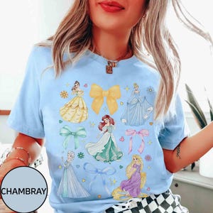 May include: Light blue t-shirt featuring illustrations of various princesses and bows. The design includes images of princesses in yellow, blue, green, and purple dresses, along with yellow, green, and pink bows. The word "CHAMBRAY" is printed on the bottom left.