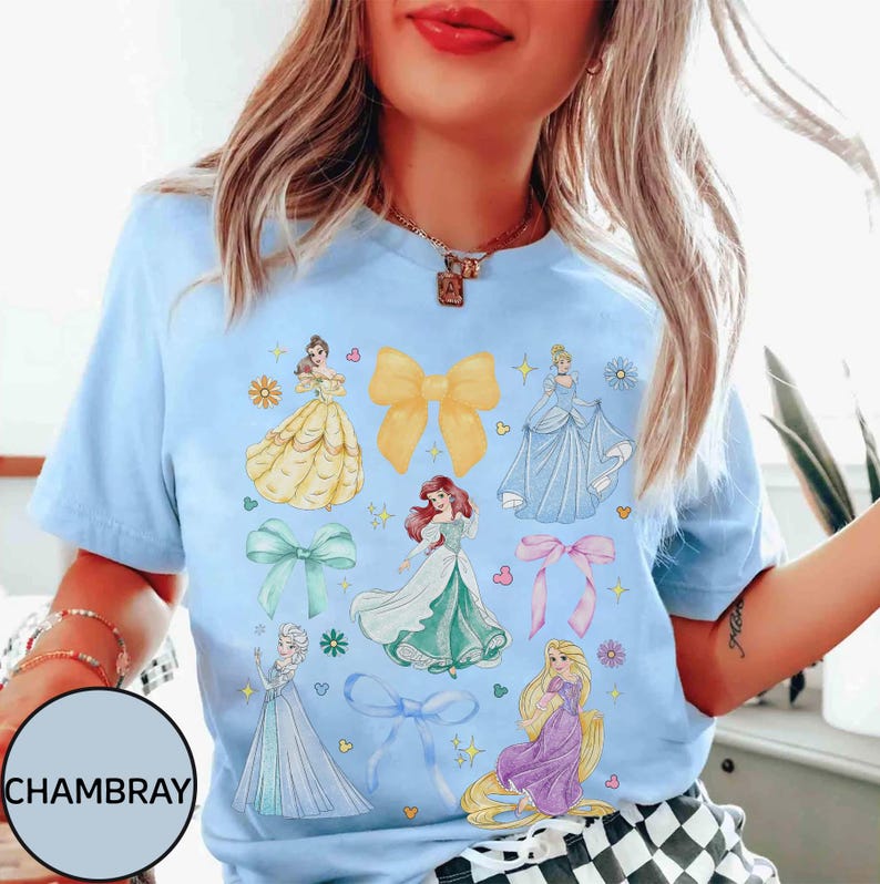 May include: Light blue t-shirt featuring illustrations of various princesses and bows. The design includes images of princesses in yellow, blue, green, and purple dresses, along with yellow, green, and pink bows. The word "CHAMBRAY" is printed on the bottom left.