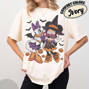 Może przedstawiać: T-shirt z kr&oacute;tkim rękawem w kolorze kości słoniowej z grafiką przedstawiającą Daisy Duck i Minnie Mouse w kostiumach na Halloween. Wz&oacute;r zawiera nietoperze, dynie oraz napis Comfort Colors Ivory. Postacie mają na sobie kapelusze czarownic.
