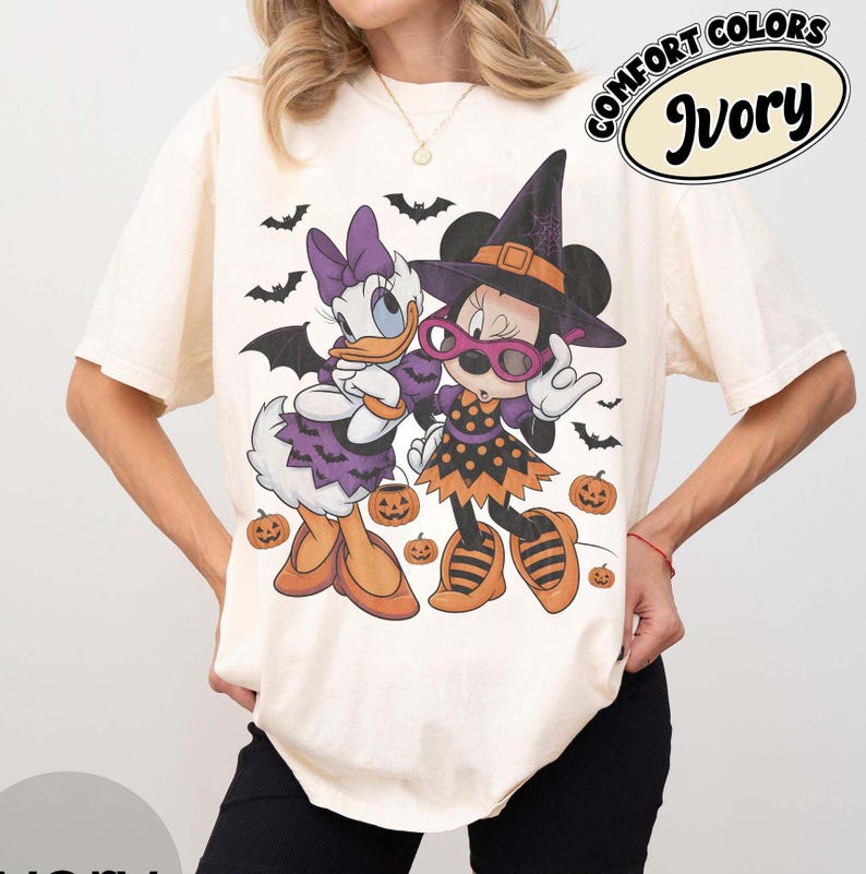 Może przedstawiać: T-shirt z kr&oacute;tkim rękawem w kolorze kości słoniowej z grafiką przedstawiającą Daisy Duck i Minnie Mouse w kostiumach na Halloween. Wz&oacute;r zawiera nietoperze, dynie oraz napis Comfort Colors Ivory. Postacie mają na sobie kapelusze czarownic.
