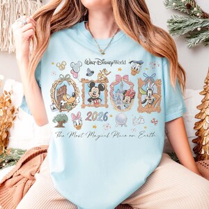 May include: Light blue t-shirt featuring framed Disney characters including Mickey Mouse, Minnie Mouse, Donald Duck, and Goofy. The shirt has the text "Walt Disney World 2026" and "The Most Magical Place on Earth."