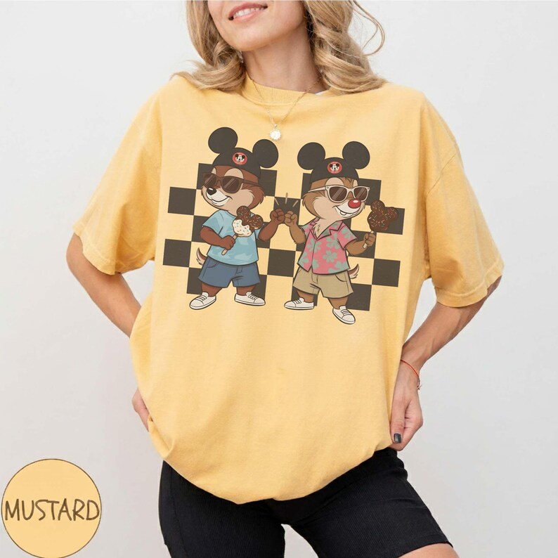 May include: Mustard yellow t-shirt featuring a graphic of two cartoon chipmunks wearing sunglasses and Mickey Mouse hats, holding ice cream cones. The chipmunks stand in front of a black and white checkered background. The word "MUSTARD" is in a circle at the bottom left.