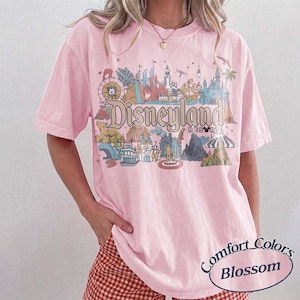 May include: A light pink t-shirt with a Disneyland Resort graphic. The graphic features various park attractions in a colorful, vintage style. The shirt also has the words "Comfort Colors Blossom" printed on it.