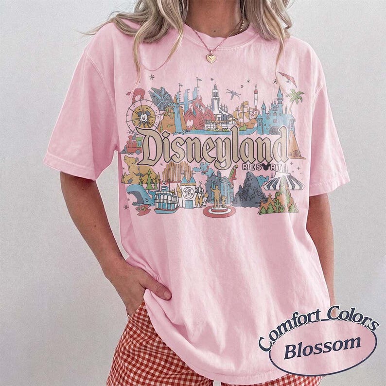 May include: A light pink t-shirt with a Disneyland Resort graphic. The graphic features various park attractions in a colorful, vintage style. The shirt also has the words "Comfort Colors Blossom" printed on it.