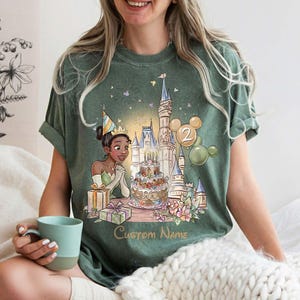 May include: Green t-shirt with a cartoon illustration of a princess celebrating a birthday with a castle, cake, and balloons. The design includes the text "Custom Name" below the illustration. The shirt is worn by a person holding a blue mug.